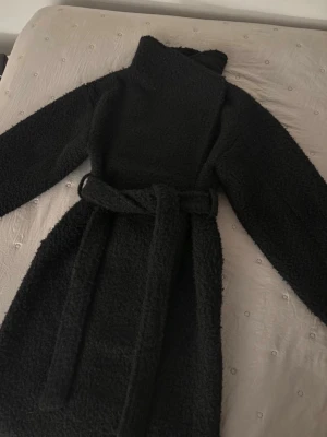 Svart lång teddyjacka med bälte - Mysig svart teddyjacka i lång modell med hög krage och matchande bälte i midjan. Jackan har en fluffig, mjuk struktur och stängs med knappar framtill. Perfekt för dig som vill ha en trendig och varm jacka med avslappnad look.
