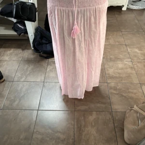 Ljusrosa långkjol från Gina Tricot - Säljer en ljusrosa långkjol från Gina Tricot med smock i midjan och dekorativ knytning med tofsar framtill. Kjolen har ett luftigt och flowy fall, perfekt för varma dagar. Använd endast några få gånger. Bara att höra av sig vid frågor🤗🫶🏻