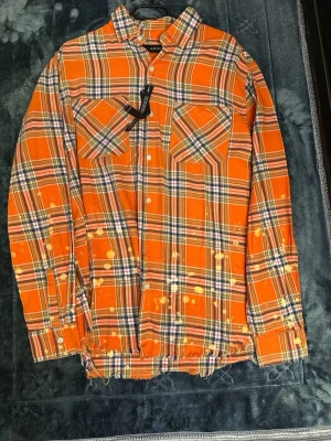  Amiri skjorta  - Säljer en orange rutig skjorta från Amiri med vita och blå detaljer. Skjortan har lång ärm, två bröstfickor, knappar framtill och slitna kanter. Stora AMIRI-bokstäver på ryggen och unika blekta detaljer för en edgy look.