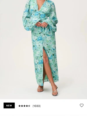 Adoore Positano dress  - Långklänning i ljusblått och grönt blommönster med v-ringad omlottdesign, långa vida ärmar och slits framtill. Klänningen har knappar längs framsidan och är tillverkad i ett lätt, flowy material som passar perfekt för sommarens alla tillfällen. Helt oanvänd, prislappen är kvar.