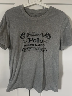 Grå t-shirt från Polo Ralph Lauren - Snygg grå t-shirt från Polo Ralph Lauren med svart tryck framtill. Klassisk rund halsringning och korta ärmar. Tillverkad i mjuk bomull som känns skön mot huden. Perfekt till jeans eller shorts för en avslappnad look.