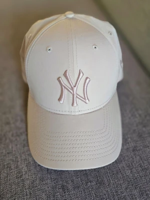 Beige New Era NY keps - Snygg beige keps från New Era med broderad NY-logga. Klassisk böjd skärm och ventilerande hål upptill. Justerbar baktill för perfekt passform. Tillverkad i ett lätt och bekvämt material som passar året runt.