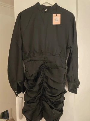 Svart ruched klänning med lång ärm - Svart kortklänning med lång ärm och hög hals. Klänningen har snygga rynkade detaljer framtill och baktill som ger en cool och trendig look. Stängs med knapp i nacken och har breda manschetter med knappar. Perfekt för dig som gillar stilrena och unika plagg. Liten i storlek! 
