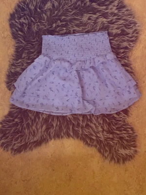 Ljusblå blommig volangkjol - Supersöt ljusblå kortkjol med blommigt mönster och volangdetaljer. Kjolen har bred smock i midjan för skön passform och är gjord i ett lätt, luftigt material som passar perfekt till varmare dagar.