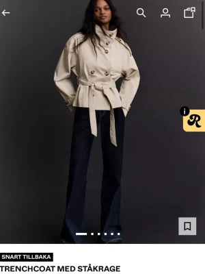 Beige trenchcoat med ståkrage - Trendig beige trenchcoat med ståkrage och dubbelknäppning. Jackan har ett knytskärp i midjan och dekorativa axelklaffar. Tillverkad i ett lätt tyg som ger en snygg siluett. Perfekt för dig som vill ha en modern och stilren look.