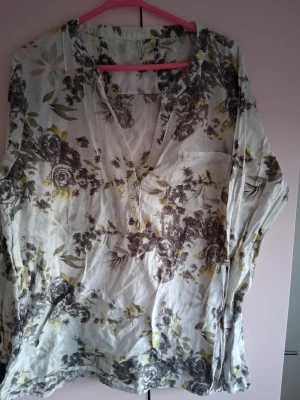 Blommig långärmad blus i vitt och grått - Säljer en luftig blus med blommigt mönster i vitt, grått och lite gult. Blusen har långa ärmar, v-ringning med knappar och en bröstficka. Perfekt till jeans eller kjol för en avslappnad look.