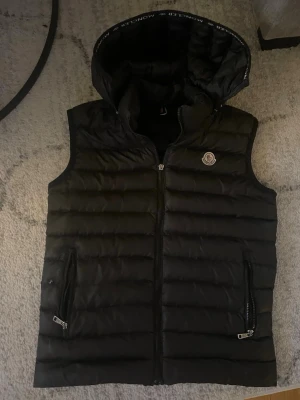 Svart dunväst från Moncler med huva - Snygg svart dunväst från Moncler med avtagbar huva och två dragkedjeförsedda fickor. Västen har Moncler-logga på bröstet och quiltad design. Perfekt för lager på lager och håller dig varm med stil. Materialet är glansigt och klassiskt för Moncler.