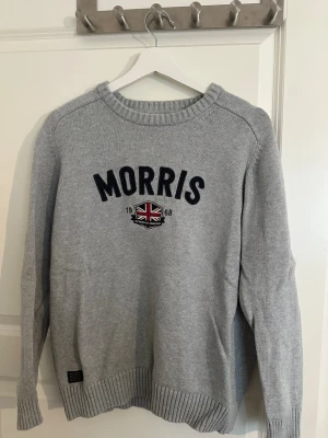 Grå stickad tröja från Morris - Snygg grå stickad tröja från Morris med rund halsringning och broderad logga samt brittisk flagga framtill. Tröjan har ribbade muddar vid ärmslut och nederkant. Perfekt för dig som gillar klassisk och stilren design.