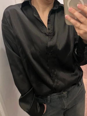 Svart satinliknande skjorta - Stilren svart skjorta i glansigt, satinliknande material. Klassisk krage, knäppning framtill och långa ärmar med manschetter. Perfekt för dig som vill ha en clean och trendig look.