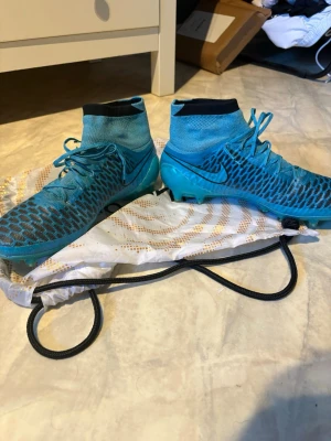 Nike Magista - Säljer ett par blå Nike fotbollsskor med svart sula och strumpkrage. Skorna har snörning, mönstrad ovandel och Nike-logga på sidan. Perfekta för dig som vill ha bra grepp och stil på planen. Materialet är syntet och textil.