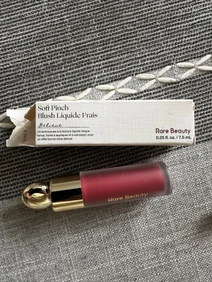 Rare Beauty Soft Pinch Liquid Blush - Flytande rouge från Rare Beauty i nyansen 'Believe', en djup rosabrun färg. Kommer i en elegant matt glasflaska med guldfärgat lock och applikator. Innehåller 7,5 ml och ger ett naturligt resultat med lätt att blenda formula.