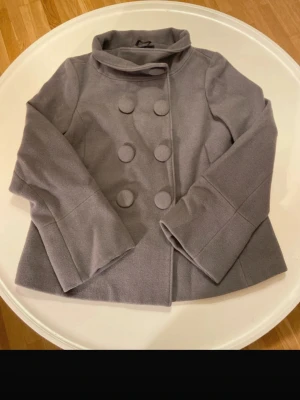 Grå dubbelknäppt rock - Pris kan diskuteras vid snabbköp. Snygg grå kappa med dubbelknäppning och stora dekorativa knappar. Modellen har en bred krage och vida ärmar som ger en trendig siluett. Tillverkad i mjukt tyg som känns skönt att bära. Perfekt för dig som vill ha en stilren och modern look. Den är lite trasig på innersidan, se bild (det är inget som syns utanpå)