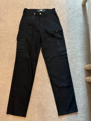 Fritidsbyxor - Svarta fritidsbyxor/arbetsbyxor. Fint skick utöver en flärp som ska vara över en ficka, men denna är bortklippt. Tycker inte det märks av när man har på sig dem.