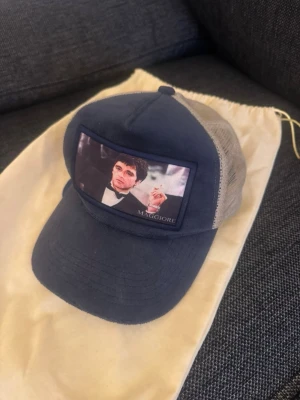 Blå truckerkeps Maggiore mesh - Säljer en blå truckerkeps från Maggiore med mesh på sidorna och baksidan. Framtill finns en patch med ett tryck av scarface och texten 'MAGGIORE'. Kepsen har böjd skärm och justerbar snapback. Perfekt för en avslappnad streetwear-look.