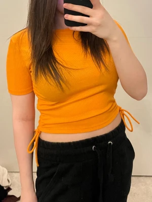 Orange ribbad croppad topp med knyt - Trendig orange ribbad topp med kort ärm och croppad passform. Snygga knytband i sidorna som ger en cool detalj. Perfekt för dig som vill sticka ut med färg! Mjuk och stretchig, passar till både jeans och kjol.