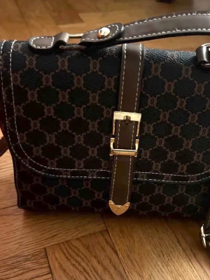  brun axelväska med monogram - Snygg brun axelväska med klassiskt monogrammönster och detaljer i mörkbrunt läder. Väskan har justerbar axelrem, guldfärgade metalldetaljer och dekorativt spänne framtill. Perfekt för dig som gillar tidlös design och lyxiga accessoarer.