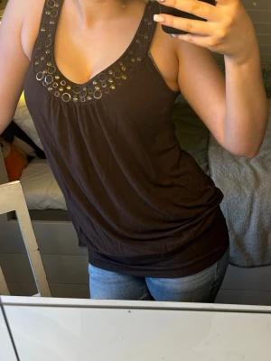 Brun linnetopp med metallringar - Snygg brun linnetopp med breda axelband och dekorativa metallringar runt urringningen. Toppen har en avslappnad passform och är perfekt för varma dagar. Tillverkad i mjukt bomullsmaterial som känns skönt mot huden. Står ingen storlek men passar mig som är en s.