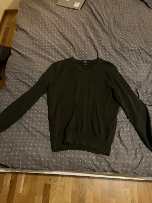 Svart stickad tröja Polo Ralph Lauren - Klassisk svart stickad tröja från Polo Ralph Lauren i mjuk pima bomull. Tröjan har v-ringning och långa ärmar, perfekt för lager på lager och enkel att matcha till olika outfits.