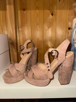 Rosa platåsandaler med chunky klack - Säljer ett par snygga rosa sandaler med bred platå och chunky klack. Skorna har korsade band framtill och justerbart spänne i silver vid ankeln. Tillverkade i mockaimitation som ger en mjuk och trendig look. Perfekta för dig som vill ha extra höjd och stil.