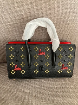 Christian Louboutin svart handväska - Snygg svart handväska från Christian Louboutin med röda detaljer och guldfärgade nitar. Väskan har ett rektangulärt format, dubbla handtag och är dekorerad med Louboutin-loggor och små metalliska detaljer. Tillverkad i strukturerat läder för en lyxig känsla.