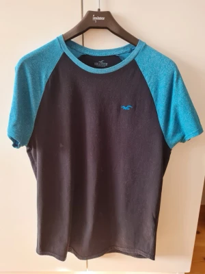 Svart och blå t-shirt från Hollister strl S - Snygg t-shirt från Hollister med svart kropp och blå ärmar. Den har en liten logga på bröstet och är kortärmad. Storlek S. Kan hämtas i Vallentuna.