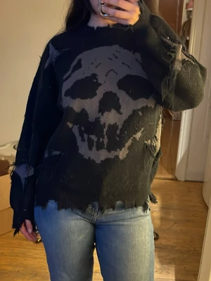 Svart stickad tröja med dödskalle - Cool svart stickad tröja med stort dödskallemotiv framtill. Tröjan har en oversized passform, slitna detaljer och råa kanter för en edgy look. Långa ärmar och mjukt material, perfekt för dig som gillar alternativ stil.