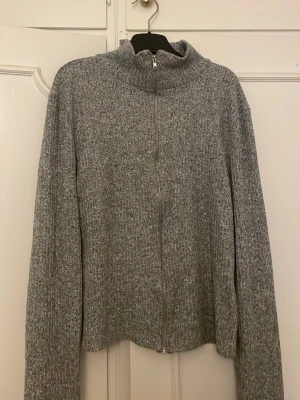 Gråmelerad stickad ziptröja - Mysig gråmelerad stickad tröja med dragkedja framtill och hög krage. Långärmad modell i mjukt material, perfekt för lager på lager. Enkel och stilren design utan synligt märke.