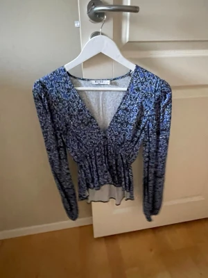 Blommig blå blus från NA-KD - Söt blommig blus från NA-KD i blått med små vita och svarta detaljer. Djup V-ringning, markerad midja och volangkant nedtill. Långärmad modell i ett lätt och luftigt tyg, perfekt för en trendig look.