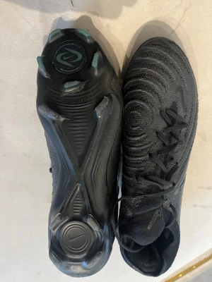 Nike luna  svarta fotbollsskor elit  - Säljer ett par helsvarta Nike fotbollsskor med högre strumpliknande skaft och snörning. Skorna har en modern design med strukturmönster på ovandelen och dobbar för spel på gräs. Tillverkade i syntetmaterial för bra grepp och kontroll.