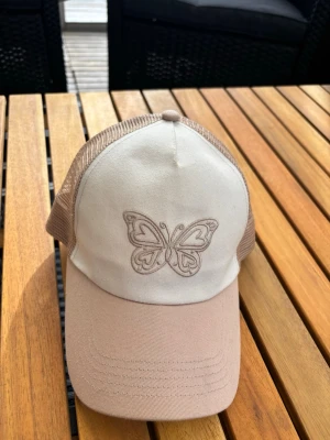 Beige truckerkeps med fjäril / y2k - Snygg beige truckerkeps med mesh på sidorna och broderad fjäril framtill. Skärmen är ljusbeige och framsidan i tyg, medan baksidan är i meshmaterial för extra ventilation. Perfekt accessoar för en chill och trendig look.