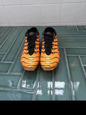Orange fotbollsskor från Nike - Säljer ett par grymma fotbollsskor från Nike i orange med svarta detaljer och coola vågformade mönster. Skorna har svarta snören och en vit sula med orange dobbar, perfekt för spel på gräs. Materialet är syntetiskt och designen är riktigt sportig.