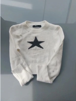 Vit stickad tröja med svart stjärna - Vit stickad tröja från new yourker  med en stor svart stjärna på bröstet. Tröjan har rund hals, långa ärmar och breda ribbade muddar vid ärmslut och nederkant. Materialet ser mjukt och mysigt ut, perfekt för kyliga dagar.