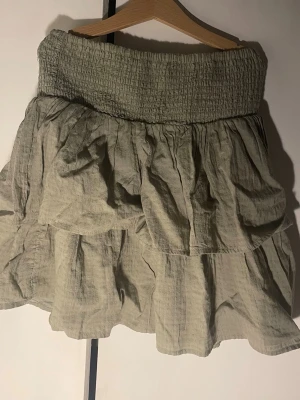 Grön volangkjol med smock - Säljer en grön kortkjol med bred smock i midjan och dubbla volanger.  Storlek 170. Andvänd kanske en gång, inget tecken på slitage. Verkligen superfin men kommer tyvärr inte till andvändning! Kjolen har en luftig och avslappnad passform, perfekt för varma dagar. Tillverkad i ett lätt bomullstyg som ger en skön känsla.