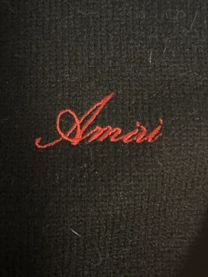 Svart hoodie från Amiri med brodyr - Svart hoodie från Amiri med röd broderad logga på bröstet och stora vita detaljer längs ena ärmen. Hoodien har huva med dragsko och känguruficka framtill. Materialet ser ut att vara mjukt och lite luddigt, perfekt för en avslappnad streetwear-look.