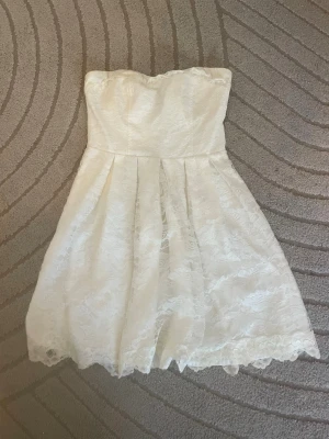 Studentklänning❤️ - Söt vit axelbandslös kortklänning med vacker spetsdetalj och lätt utställd kjol. Perfekt för sommarens alla tillfällen. Klänningen har en feminin siluett och romantisk känsla tack vare spetsen. Inbyggd byggel