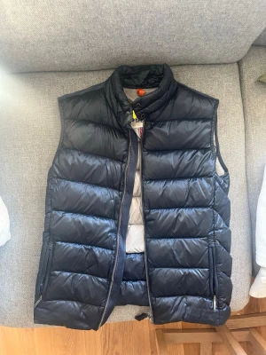 Svart dunväst från Parajumpers - Snygg svart dunväst från Parajumpers med hög krage, dragkedja och två fickor med dragkedja. Klassisk quiltad design och ikonisk gul detalj vid halsen. Perfekt för lager-på-lager och håller dig varm under kyliga dagar. Storlek youth L sitter som en s/m 