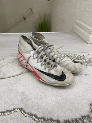Nike vita fotbollsskor med röd detalj - Säljer ett par vita Nike fotbollsskor med högt skaft och svarta swoosh-loggor. Skorna har röda detaljer på sidorna och är designade för bra grepp på planen. Snygg och modern look, perfekta för dig som vill sticka ut på matchen.