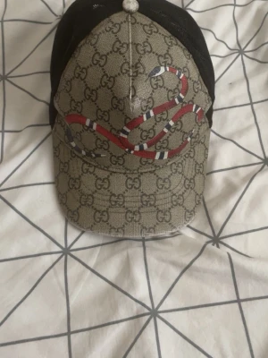 Gucci keps med orm och GG-mönster - Snygg keps från Gucci med klassiskt GG-monogram i beige och brunt. Framsidan pryds av en röd, vit och svart orm. Baksidan är i svart mesh för extra ventilation. Perfekt accessoar för en trendig streetwear-look.