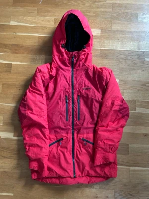 Röd vindjacka från Eqpe med huva - Snygg röd vindjacka från Eqpe med justerbar huva och dragkedja framtill. Jackan har fyra praktiska fickor med dragkedja och svart foder i huvan. Perfekt för blåsiga dagar och outdoor-äventyr. Materialet är lätt och vindtätt.