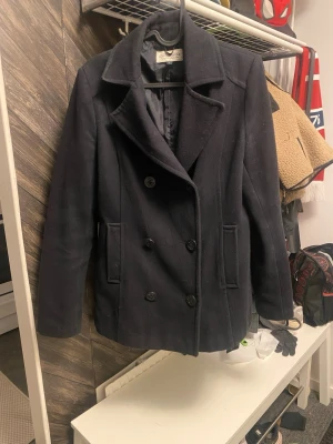 Marinblå skepparkavaj Hampton Republic - Snygg marinblå skepparkavaj från Hampton Republic med klassisk dubbelknäppning och breda slag. Jackan har två framfickor och är fodrad med svart tyg. Perfekt för dig som vill ha en stilren och tidlös look. Färgen är marinblå/svart.  Jätte fint skick på jackan! 