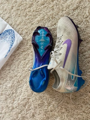 Nike Mercurial fotbollsskor vit/lilaElite  - Säljer ett par Nike Mercurial fotbollsskor elite i vitt med lila och blå detaljer. Skorna har en modern, strömlinjeformad design med syntetiskt material och en sula i blått och lila för extra grepp på planen. Perfekta för dig som vill ha stil och prestanda på fotbollsplanen.