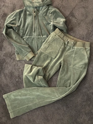 Grön velourset från Juicy Couture - Mysigt loungewear-set i grön velour från Juicy Couture. Setet består av en hoodie med dragkedja och fickor samt matchande byxor med resårmidja och dragsko. Båda delarna har diskret Juicy Couture-logga. Perfekt för chill dagar hemma.