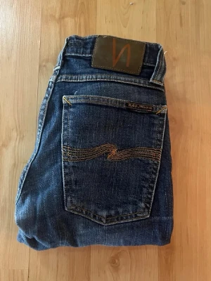 Nudie jeans  - Mörkblå jeans från Nudie Jeans, tajt long John. Midja 32cm, längd 99cm. OBS jeansen har hål på ena låret och vid knäna - se bild. JAG SKICKAR EJ FLER BILDER NÄR JAG BÄR JEANSEN! för bättre inblick i passformen rekommenderar jag att googla på modellnamnet. För storleksguide kolla måtten noggrant då jeansen alltid kan vara uppsydda heller krympta och jag ej tar emot returer :) s6,3