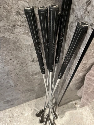 Golfklubbor PING järnset 5-9 + Wedge - Säljer ett set med sex stycken golfklubbor från PING, järn 5, 6, 7, 8, 9 samt wedge. Klubborna har svarta grepp med mönster och silverfärgade skaft i metall. Perfekt för dig som vill ha ett komplett järnset till golfbanan.