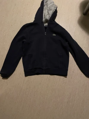 Navy zip up Lacoste - Snygg navy zip up hoodie från Lacoste med dragkedja framtill och grått innerfoder i huvan. Klassisk krokodil-logga på bröstet och två fickor på sidorna. Perfekt för en chill och stilren look. Tillverkad i mjukt bomullsmaterial.