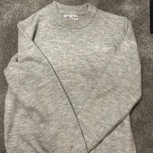 Ljusgrå stickad tröja Amé Doré - Ljusgrå stickad tröja från Aimé Doré med rund halsringning och diskret broderad logga på bröstet. Tröjan har en klassisk passform och ribbstickade muddar vid ärmslut och nederkant. Perfekt för lager-på-lager och enkel att matcha.