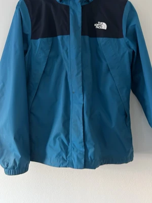 Blå vindjacka från The North Face - Snygg blå vindjacka från The North Face med svarta axelpartier och vit logga på bröstet. Jackan har dragkedja framtill, elastiska muddar vid ärmsluten och är tillverkad i ett lätt, vindtätt material. Perfekt för blåsiga dagar och outdoor-äventyr. Köptes ny från Inter sport knappt använd. Pris ej hugget i sten 🪨 👍