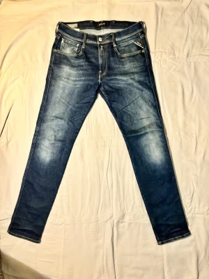 Mörkblå Replay Anbass slim fit jeans med slitningar - Snygga mörkblå slim fit jeans från märket Replay/model Anbass/ med diskreta slitningar och ljusare partier på låren. Klassisk femficksmodell med kontrastsömmar och dragkedjegylf. Perfekta för en trendig och avslappnad look. Pyttelitet hål vid knäet (se sista bild)