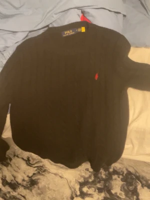 Svart kabelstickad tröja Polo Ralph Lauren - Kabelstickad svart tröja från Polo Ralph Lauren med klassisk röd logga broderad på bröstet. Tröjan har rund halsringning och ribbade muddar vid ärmslut och nederkant. Perfekt för dig som gillar stilrena och tidlösa plagg.