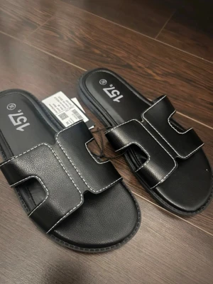 Svarta slip-in sandaler från 157 - Stilrena svarta slip-in sandaler från 157 med breda remmar i konstläder och vita kontrastsömmar. Öppen tå och platt sula för en avslappnad look. Perfekta för sommaren och enkla att matcha med olika outfits.